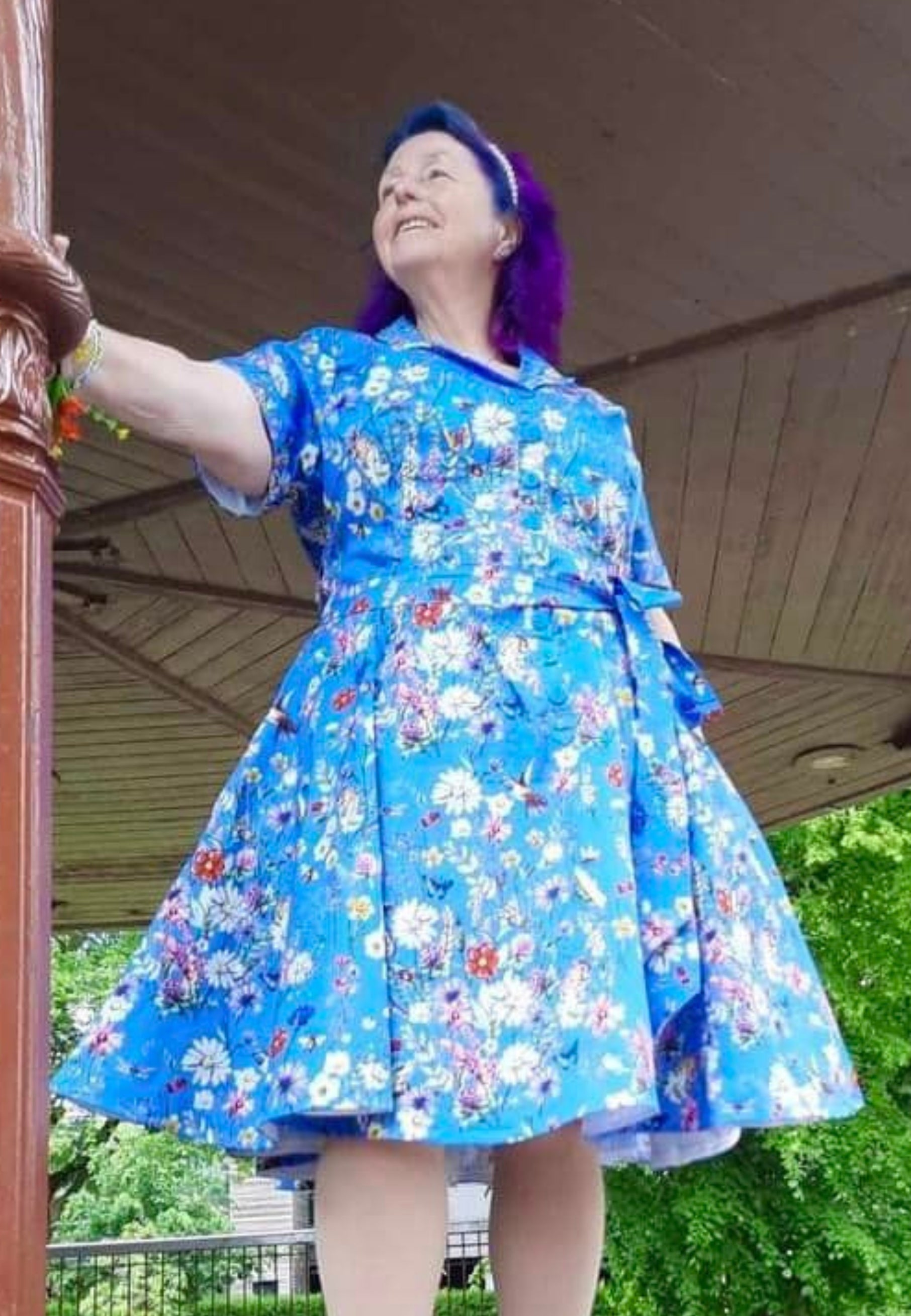 Woman in a beautiful blue retro full skirted dress with pockets and a belt. Print on the dress is a wonderful deep cornflower blue with illustrations of wild meadow flowers and insects, frogs, mice. 