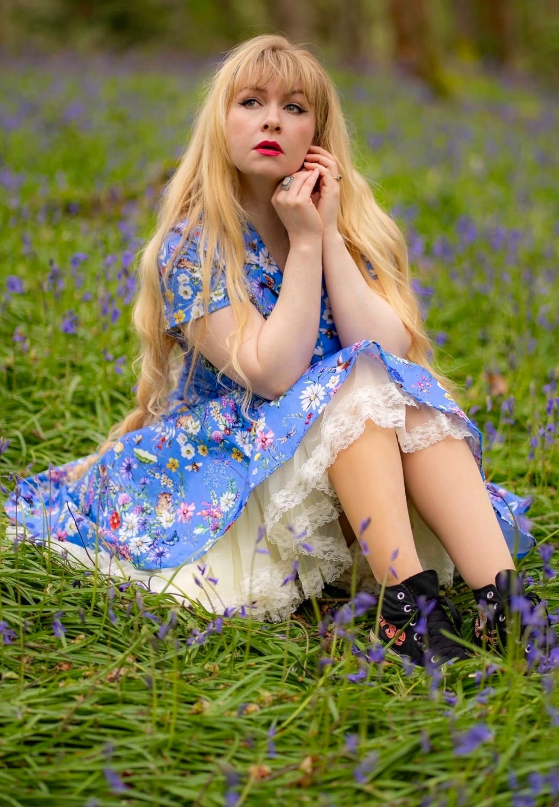 Woman with long blonde hair sat in a wild flower area of a wood wearing a beautiful cornflower blue dress with a wild flower print. 