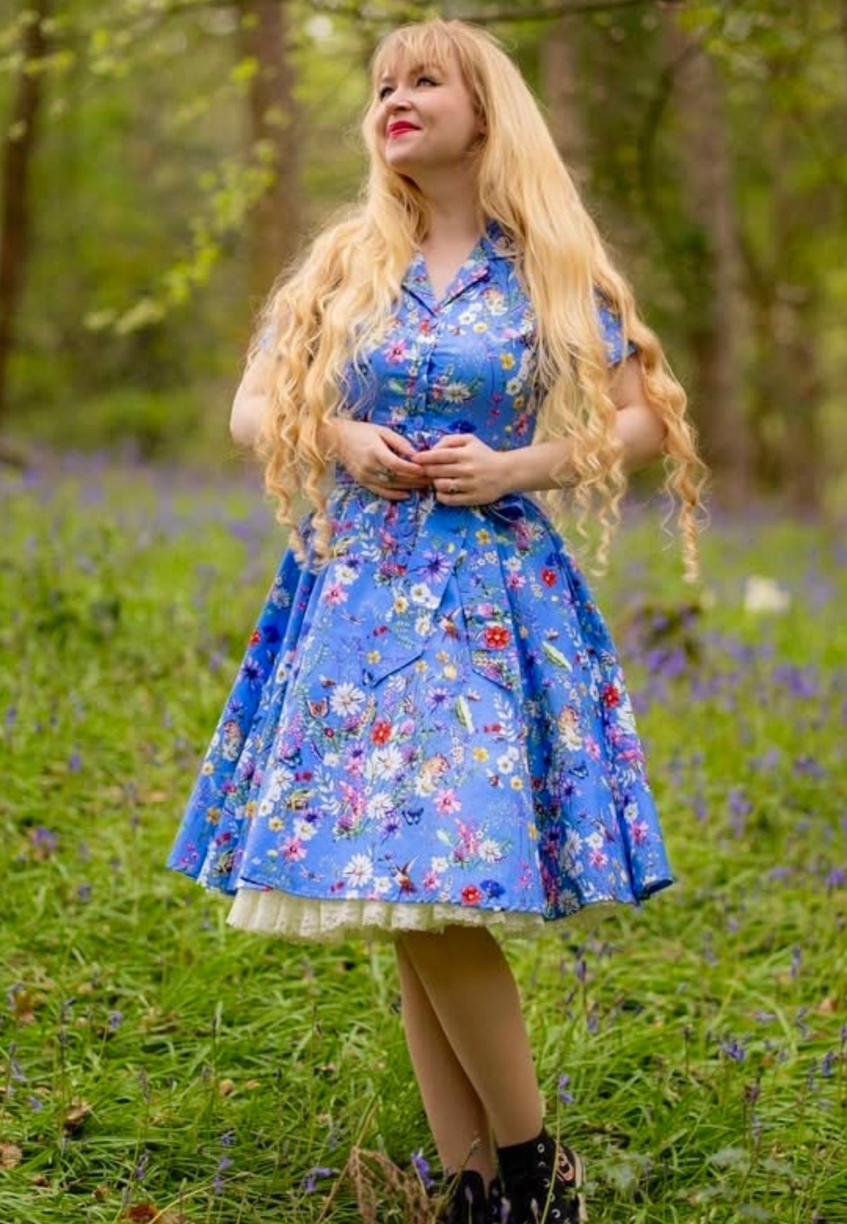 Woman with long blonde hair stood in a wild flower area of a wood wearing a beautiful cornflower blue dress with a wild flower print. 