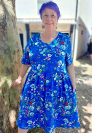 A woman with purple hair wearing a bright blue short-sleeve dress with a beautiful wild meadows print. Woman is standing outdoors next to a tree trunk.