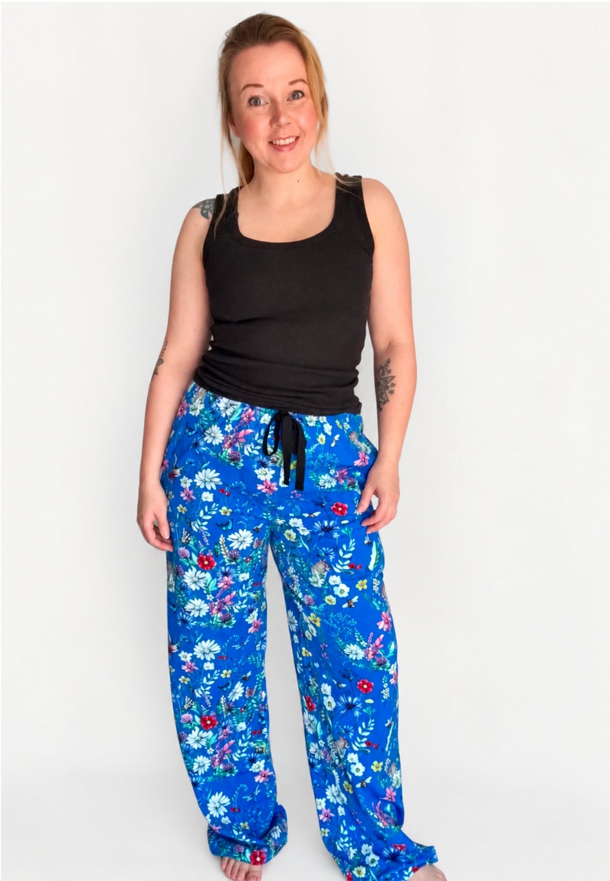 A woman modelling a pair of blue wide leg lounge pants with a beautiful wild meadows print, paired with a black tank top, against a plain white background.