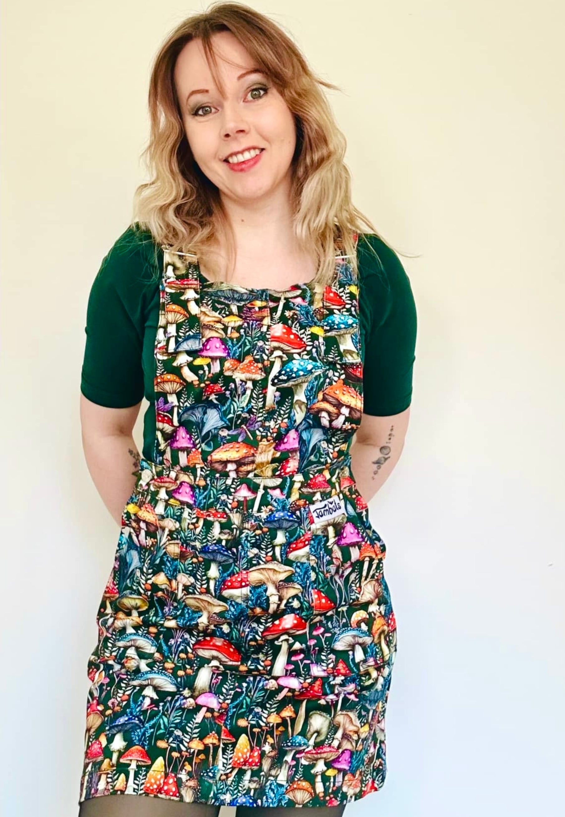A woman is wearing a dark green pinafore dress with a vibrant mushroom print. Studio photo on a plain white background. 