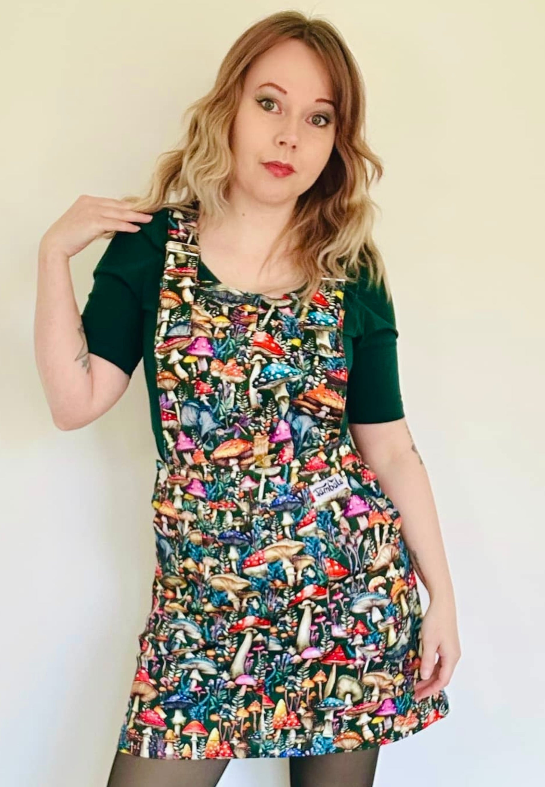 A woman wearing a dark green pinafore dress with a vibrant mushroom print with mushrooms and toadstools. Studio photo on a plain white background. 