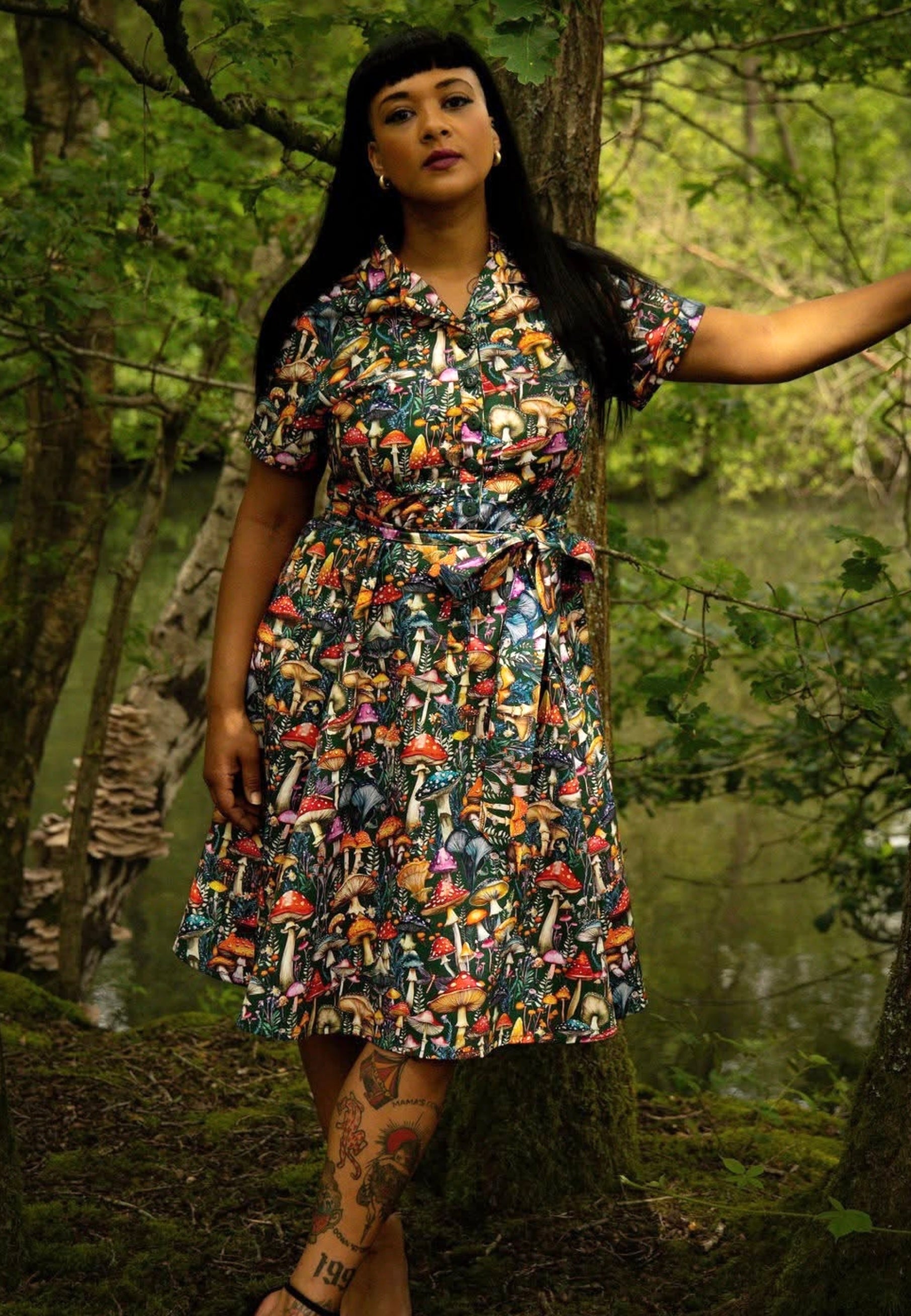 A woman wearing a short-sleeve button-up dress with a colourful retro mushroom print, standing in a forest next to a tree trunk.