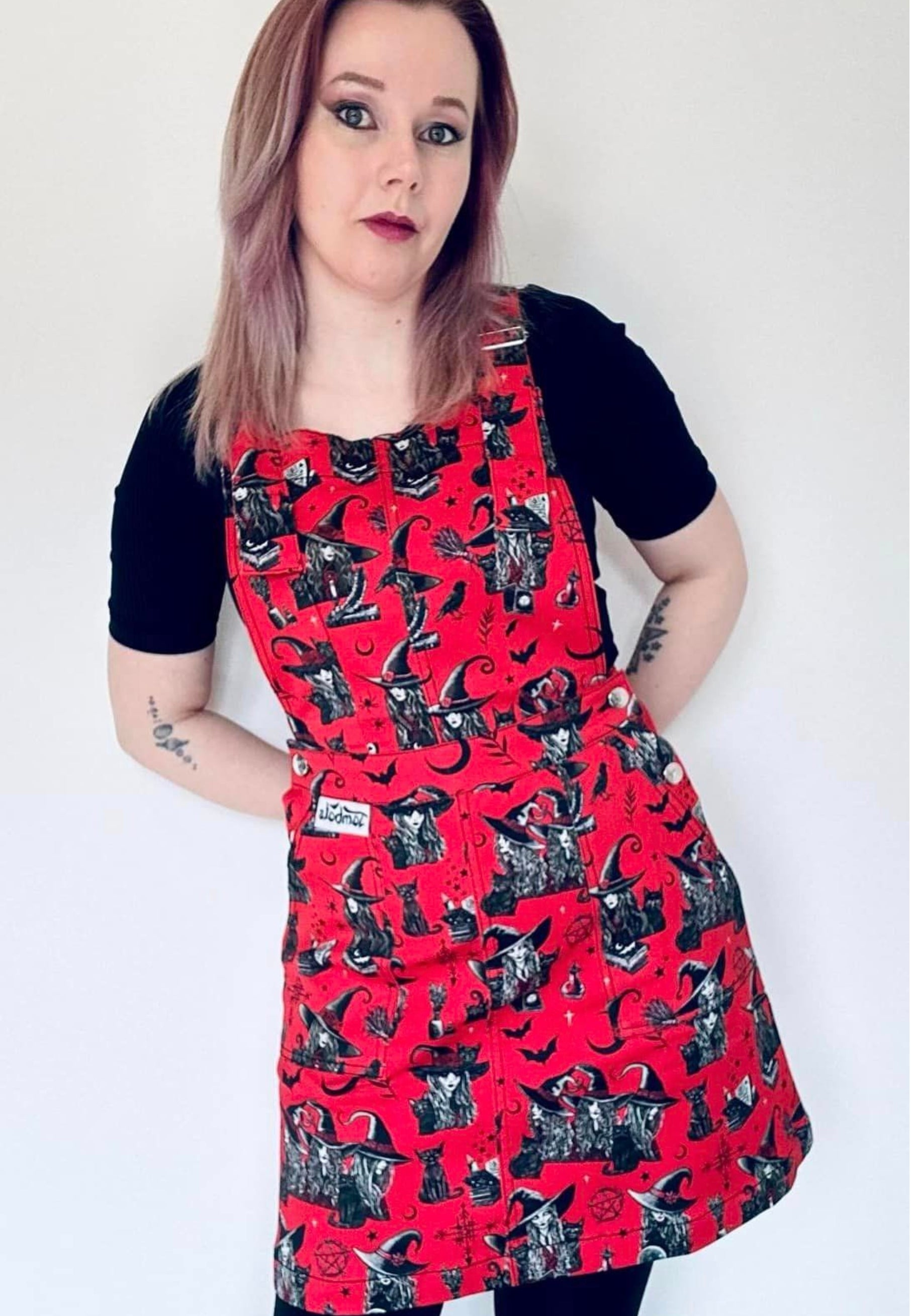 Model wearing a red pinafore dress with a witchy print, featuring deep hip pockets and side button fastenings.