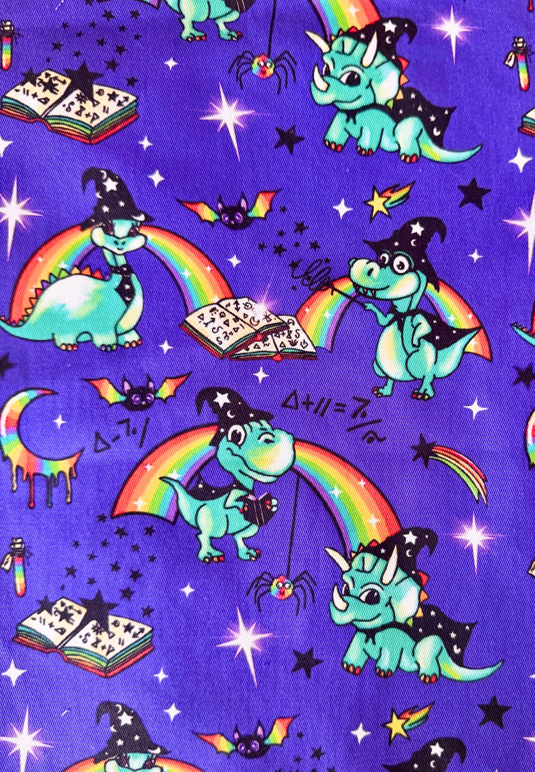A repeating fabric pattern on a purple background featuring wizard dinosaurs, rainbows, stars, bats, moons, and spells in bright colors.