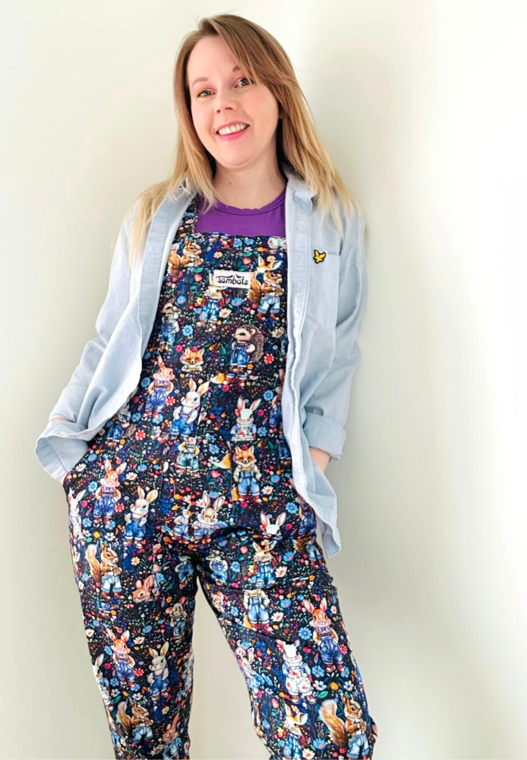 Woman wearing a pair of multicoloured cotton dungarees with a unique print of whimsical woodland creatures like a rabbit, fox, hedgehog, mole..all wearing denim dungarees on a floral background. Paired with a denim jacket.  