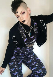 A person wearing black dungarees with a unique hand illustrated bat in space print, featuring multiple pockets and slider buckle fasteners.