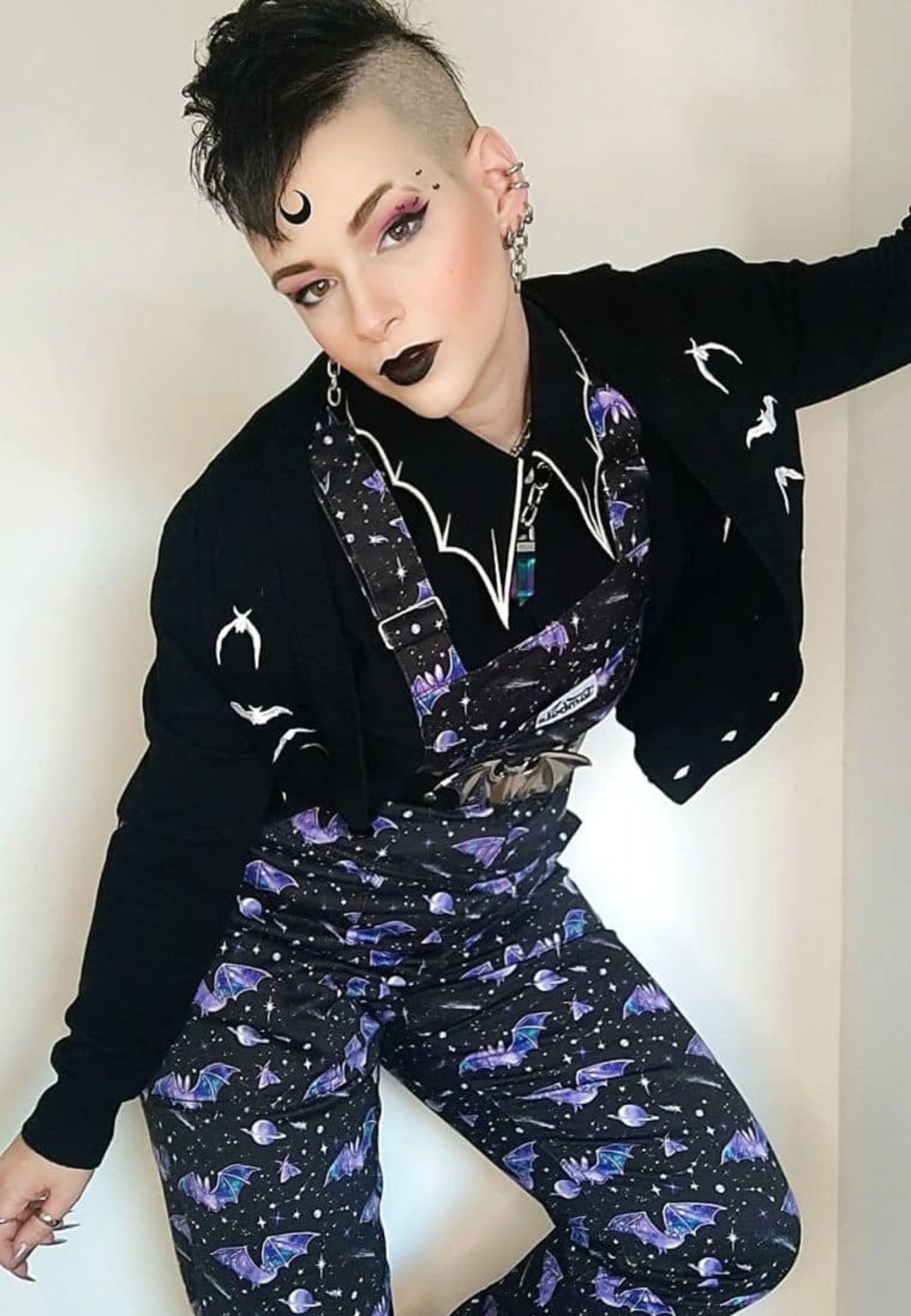 A person wearing black dungarees with a unique hand illustrated bat in space print, featuring multiple pockets and slider buckle fasteners.