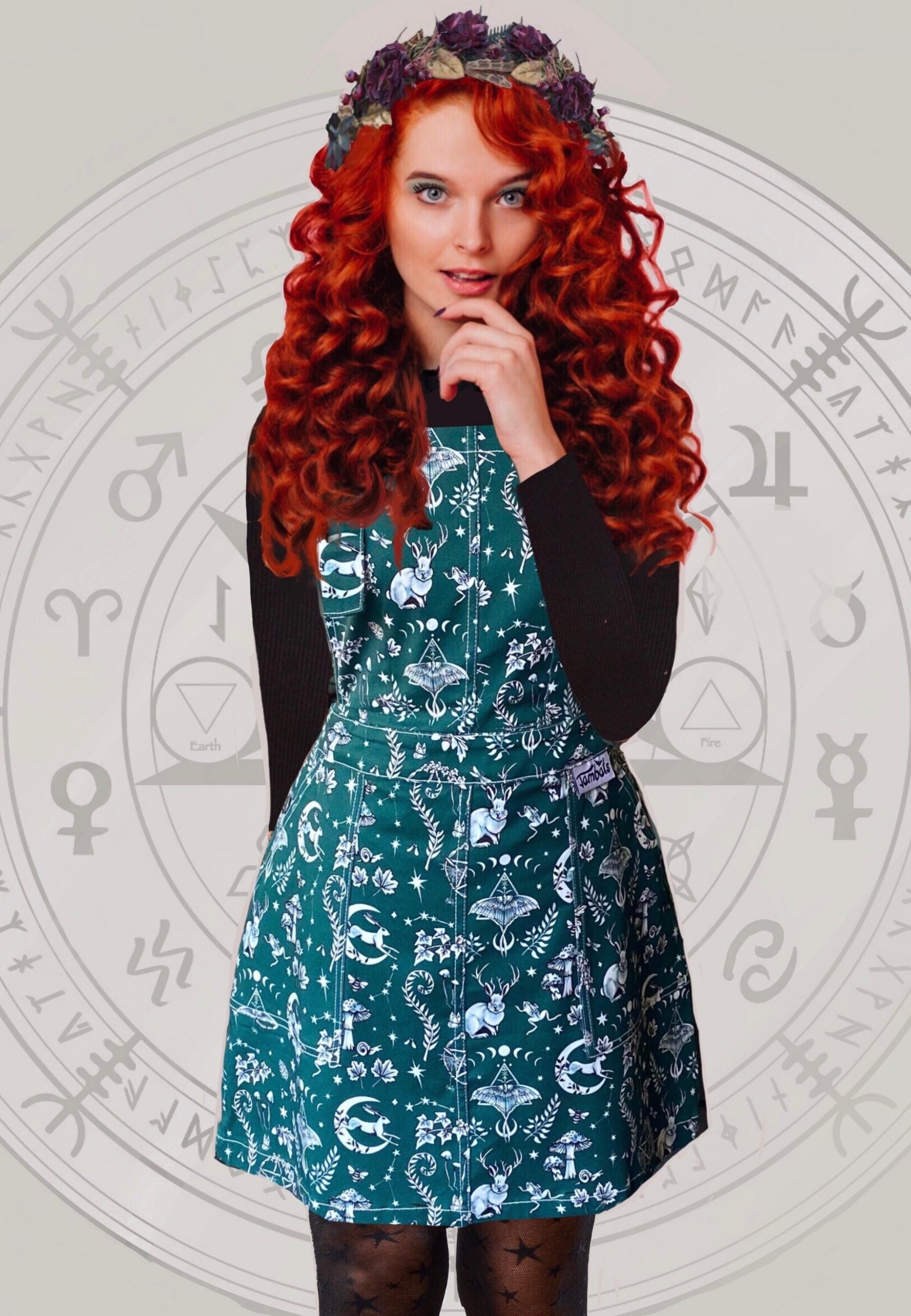 A woman with long red hair wearing a deep green pinafore dress with a white Magick print, including moon and hare, luna moth, and other magical elements. The dress features deep hip pockets and side button fastenings.